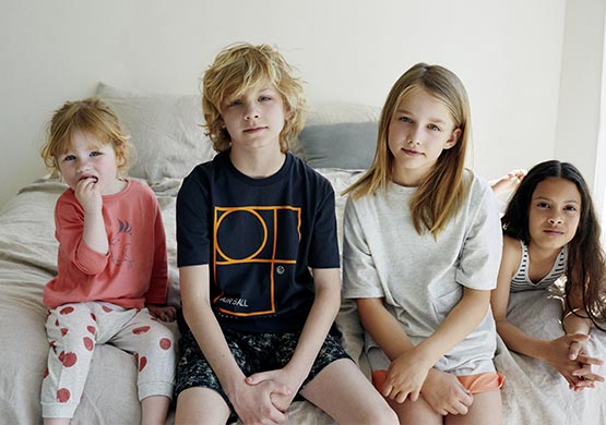 Vier Kinder mit Kinderbekleidung sitzen auf der Bettkante.