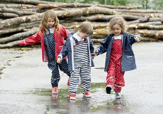 Drei Kinder mit Regenbekleidung laufen durch Pfützen.