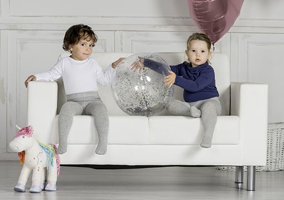 Zwei Kinder auf einer Kindercouch mit Strumpfhosen und Oberteilen bekleidet, spielen mit einem Ball.